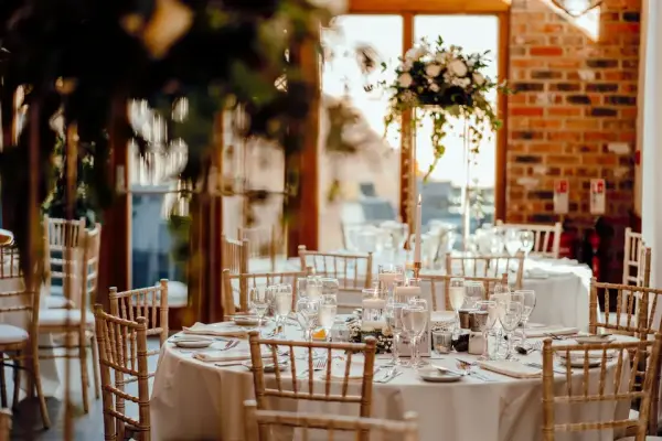 Elegant dining setup with round tables, crystal glassware, and floral centerpieces, against a backdrop of warm brick and natural light.