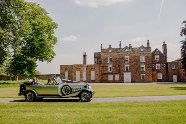 A vintage car drives past a charming brick venue, surrounded by lush green lawns and tall trees, perfect for outdoor celebrations.