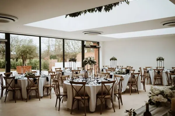Bright event space featuring elegant round tables with floral centerpieces, surrounded by large windows and natural greenery. Ideal for weddings.