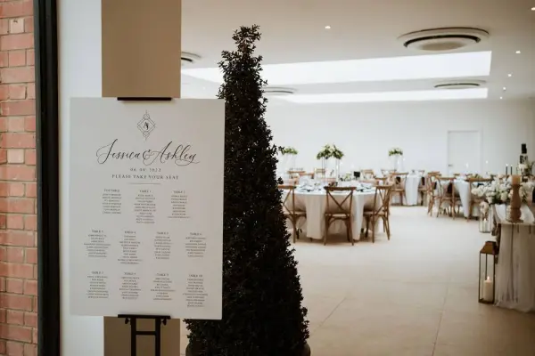 Bright reception area featuring elegant round tables with floral centerpieces, a welcome sign, and greenery accents for a wedding atmosphere.
