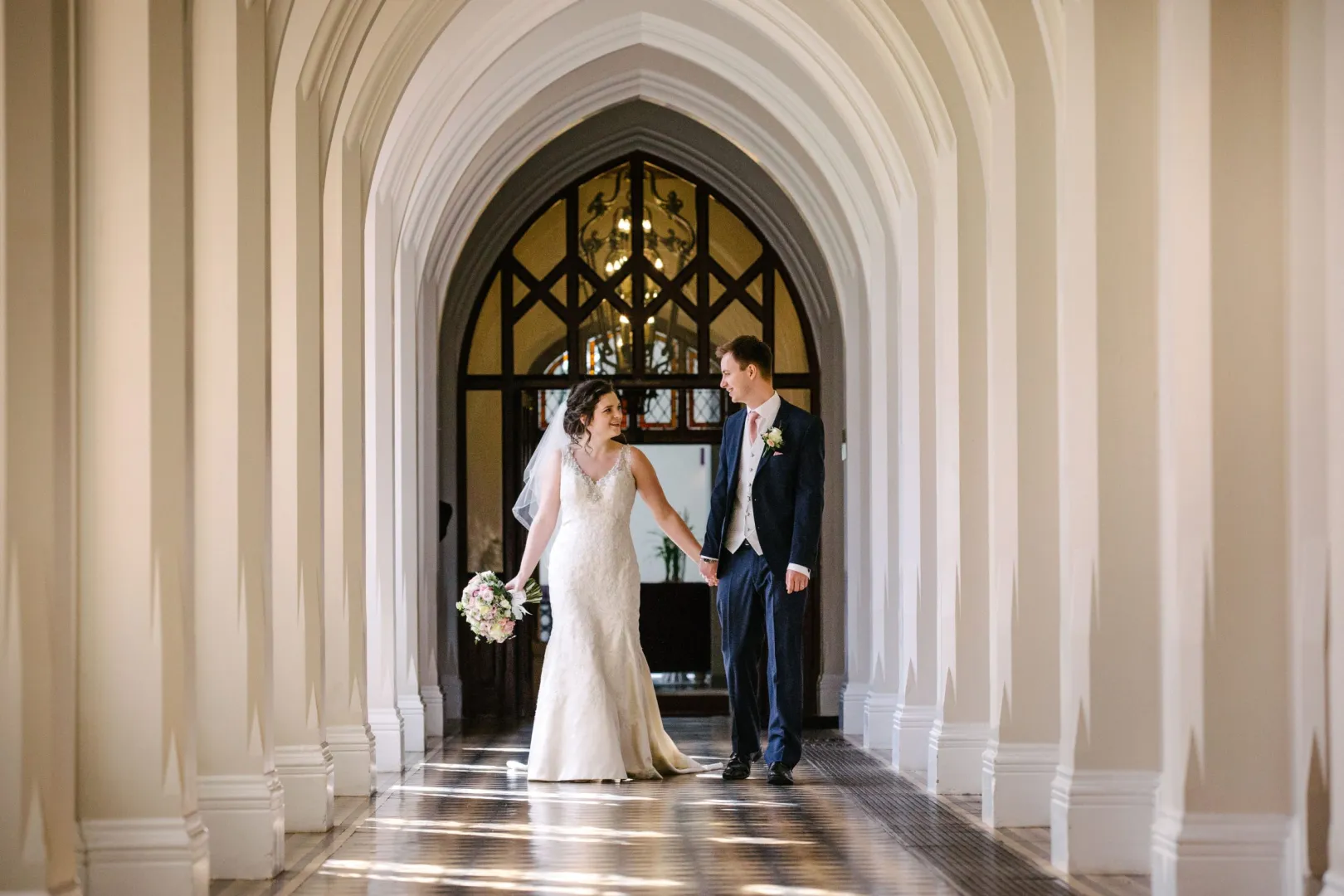 Couple walking hand-in-hand through an elegant arched hallway, adorned with soft lighting and a stunning chandelier, perfect for wedding photos.