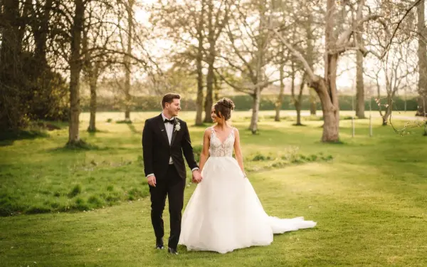 A bride in a flowing gown and a groom in a tuxedo stroll hand-in-hand through a lush, green garden dotted with trees.