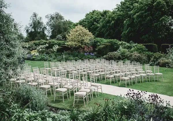 A lush garden venue with neatly arranged beige chairs for an outdoor wedding ceremony, surrounded by vibrant greenery and colorful blooms.