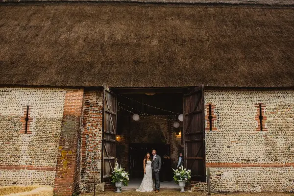 Charming rustic barn entrance showcases a couple under soft lighting, framed by large wooden doors and elegant floral arrangements.