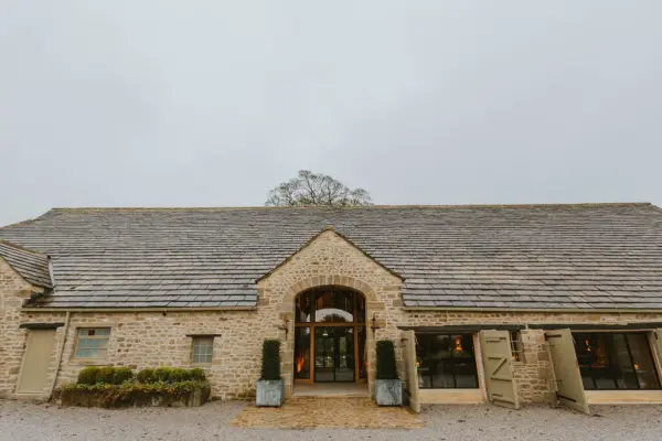 Charming rustic barn with a stone exterior and large entrance doors, surrounded by greenery and a gravel pathway, perfect for weddings.