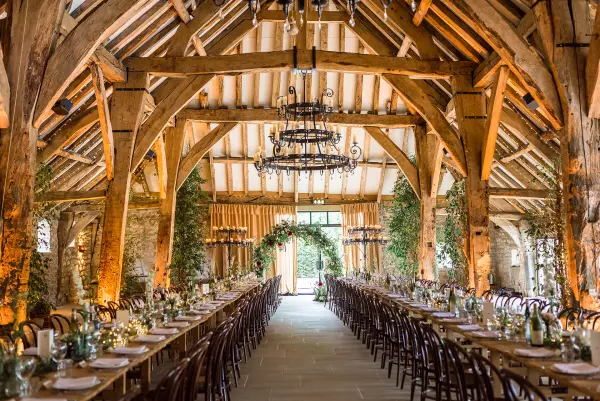 Rustic barn venue featuring exposed wooden beams, chandeliers, and long banquet tables adorned with greenery for a charming reception.