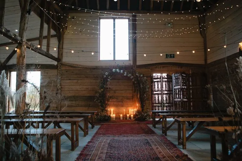 A rustic barn venue featuring wooden beams, twinkling fairy lights, a floral arch, and a cozy aisle with candles, perfect for intimate ceremonies.