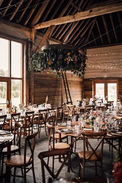 Rustic barn reception space featuring wooden beams, round tables, elegant centerpieces, and a vibrant floral chandelier. Natural light flows in through large windows.