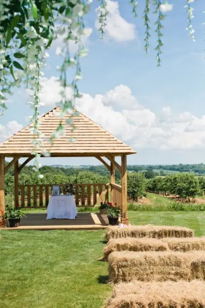 Charming outdoor wedding ceremony site featuring a rustic wooden gazebo, hay bales for seating, and scenic vineyard views.