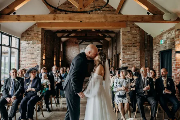 Intimate wedding ceremony in a rustic venue with exposed brick walls, large windows, and wooden beams, capturing a heartfelt kiss.