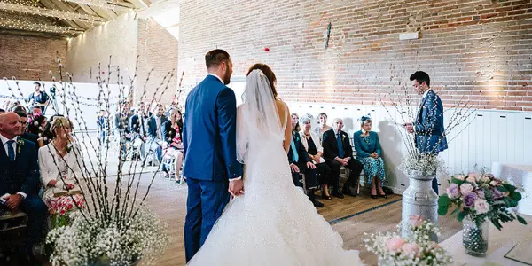 Rustic ceremony space with exposed brick walls, elegant floral arrangements, and seated guests, framing the couple during their vows.