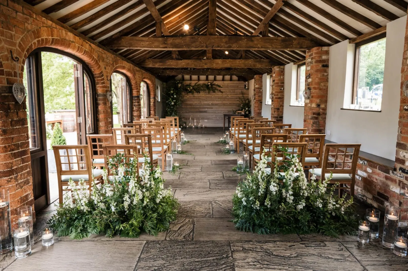 Charming rustic ceremony space featuring exposed beams, brick walls, and floral arrangements framing elegant wooden chairs.