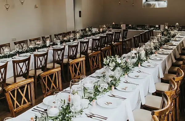 Long banquet tables adorned with lush greenery centerpieces, elegant settings, and warm wooden chairs create a charming dining atmosphere.
