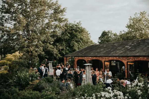 Charming outdoor wedding reception featuring guests mingling in a lush garden, with a cozy brick venue and string lights adorning the space.