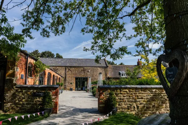 Charming rustic barn venue entrance with stone walls, lush greenery, and a pathway leading to a vibrant outdoor ceremony space.