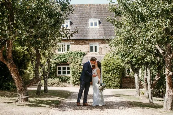 A couple shares a romantic kiss under lush trees, with a charming stone cottage in the background, ideal for intimate outdoor weddings.