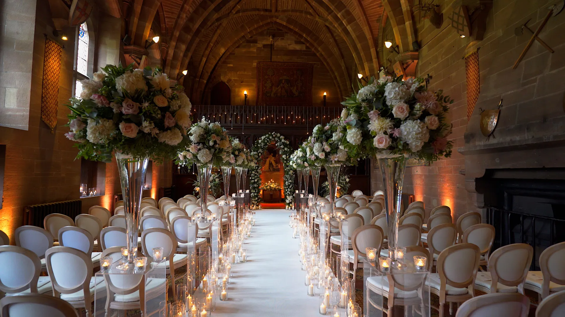Elegant indoor ceremony space featuring tall floral arrangements, white chairs, and glowing candles along a soft aisle. Stunning vaulted ceiling enhances charm.