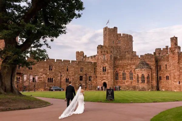 Couple walks hand in hand toward a historic stone castle, featuring lush green lawns and majestic towers, perfect for a romantic wedding backdrop.