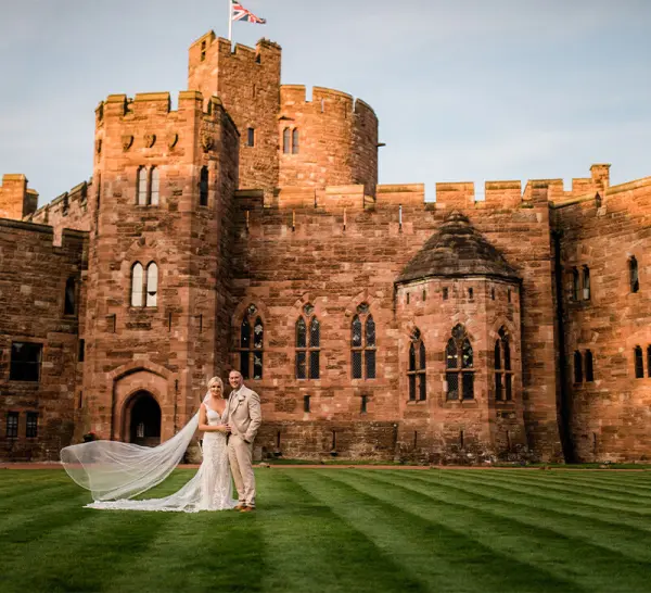 A bride and groom stand on lush green grass in front of a majestic stone castle, showcasing stunning architecture and historic charm.