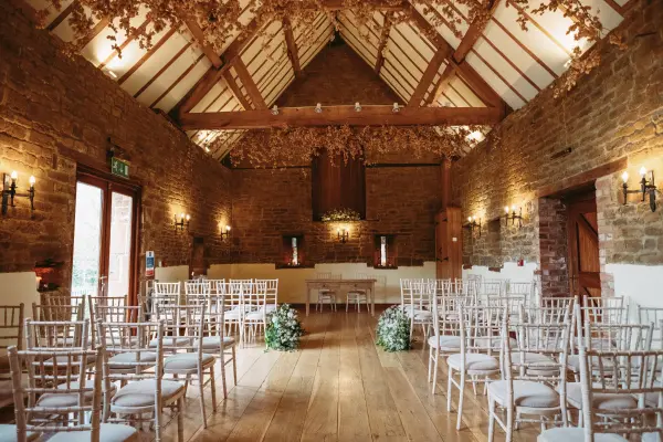 Rustic ceremony space featuring wooden beams, stone walls, and elegant chiavari chairs adorned with greenery for a charming celebration.