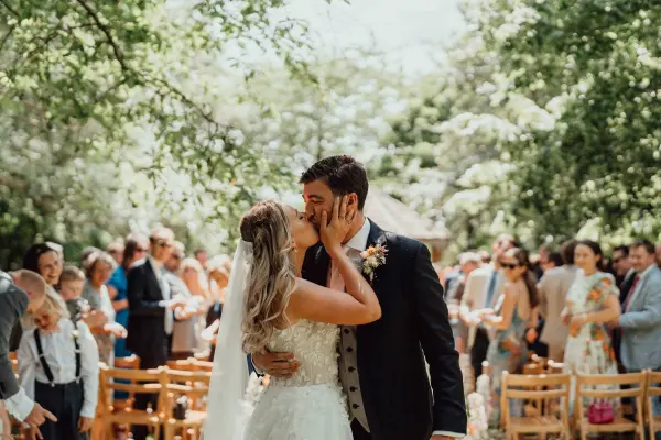 A joyful couple kisses amid lush greenery, surrounded by seated guests in a charming outdoor ceremony space, creating a romantic ambiance.