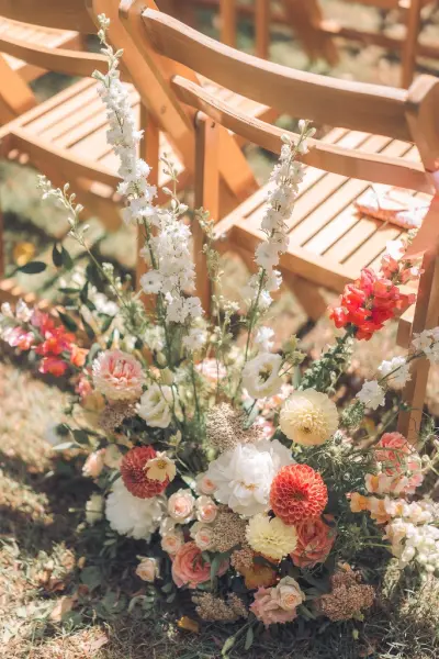 An elegant floral arrangement featuring soft pastel blooms beside wooden ceremony chairs, creating a romantic outdoor wedding ambiance.