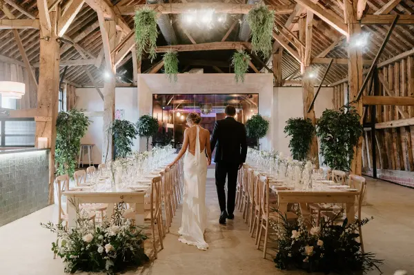 Couple walking down an aisle in a rustic barn, featuring elegant wooden beams, lush greenery, and intimate seating arrangements.