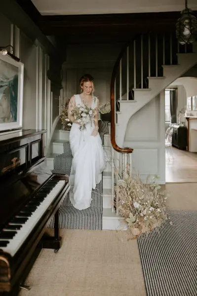 A bride descends a staircase, holding a bouquet, with a chic piano and elegant decor enhancing the venue's timeless charm.