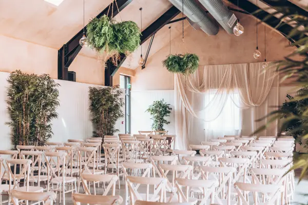 An airy indoor ceremony space featuring rows of white wooden chairs, lush greenery, and elegant drapery for a romantic atmosphere.