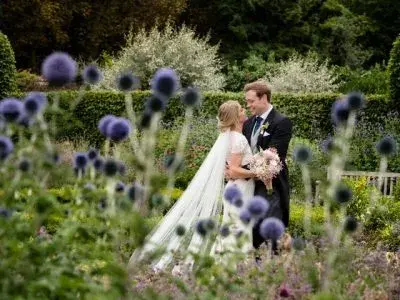 A couple embraces in a blossoming garden, framed by lush greenery and vibrant purple flowers, creating a romantic outdoor setting.