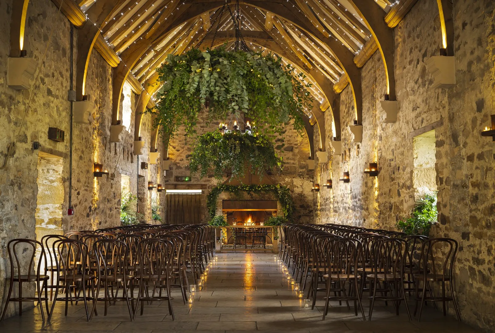 Charming stone venue featuring a high ceiling with wooden beams, lush greenery overhead, and rows of elegant chairs lining a warm aisle.