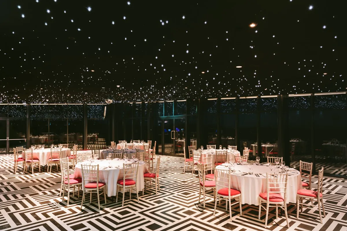 Elegant wedding reception space featuring round tables with white linens and pink accents under a starry black ceiling and geometric floor design.