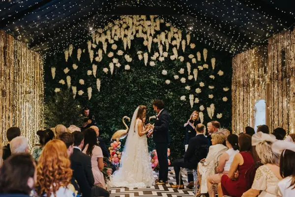 Intimate wedding ceremony featuring a lush greenery backdrop, suspended floral decor, and twinkling fairy lights in a stylish indoor setting.