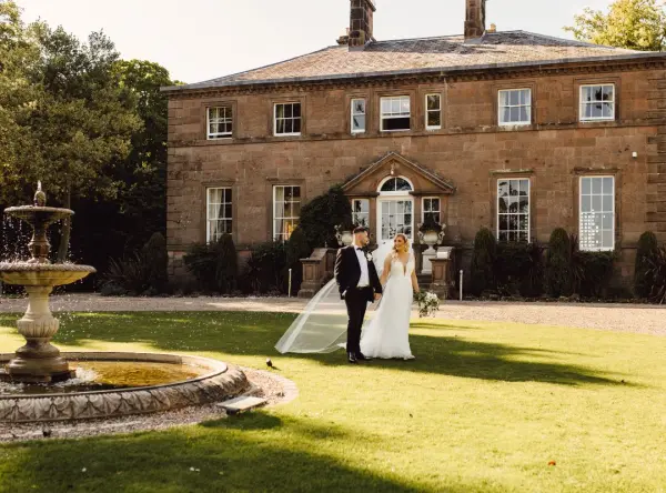 Couple walks hand-in-hand on the lush lawn, with an elegant historic mansion and fountain creating a romantic wedding backdrop.