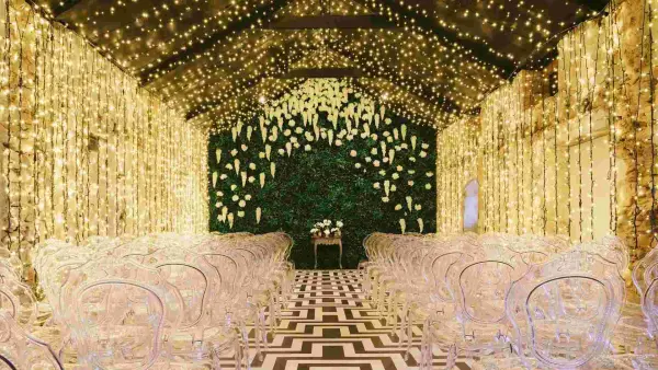 A stunning indoor ceremony space with string lights, a lush greenery backdrop, and elegant clear chairs on a patterned floor.