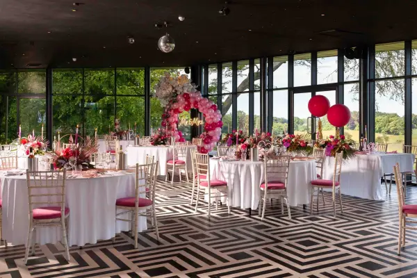 A modern reception space with elegant tables adorned in white linens, pink chairs, and vibrant floral arrangements, featuring large windows for natural light.