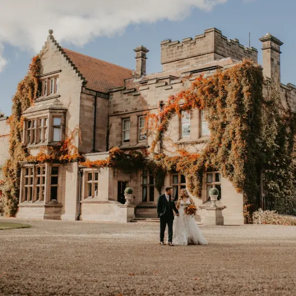 A couple stands together in front of a charming stone estate adorned with vibrant autumn foliage, creating a romantic backdrop.