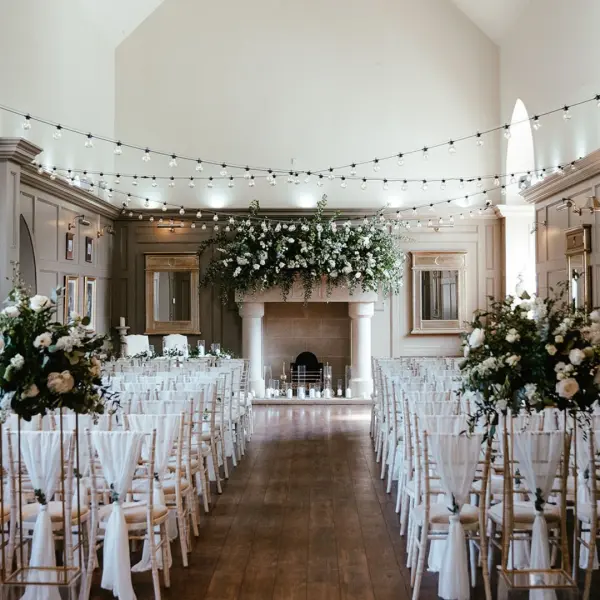 Elegant indoor ceremony space featuring a fireplace, floral arrangements, and string lights, with rows of white chairs arranged for guests.