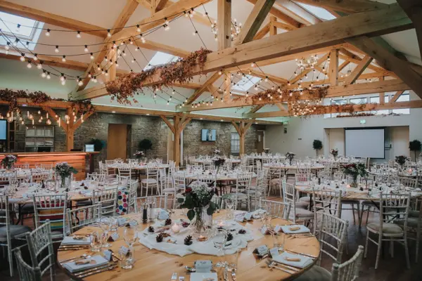 Stylish reception space featuring rustic beams, fairy lights, elegant tables set with floral centerpieces, and a warm, inviting atmosphere.