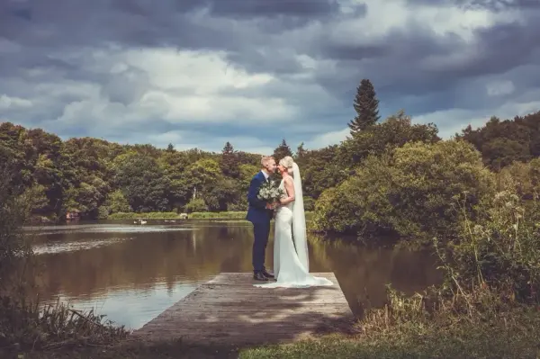 A couple shares a romantic kiss on a wooden dock by a scenic lake, surrounded by lush greenery and dramatic clouds above.