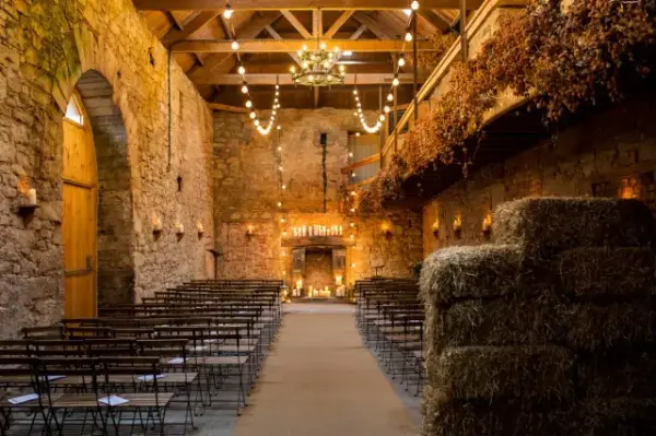 Rustic stone barn with exposed beams, chic chandelier, and hay bales, set up for a cozy wedding ceremony with string lights and guest seating.