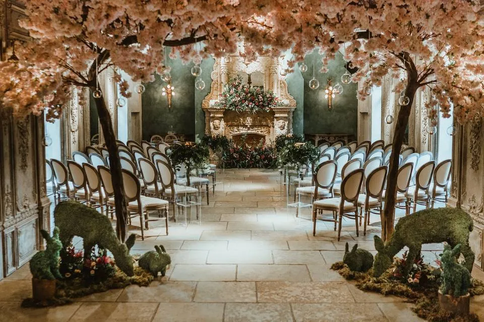 A charming indoor ceremony space adorned with pink blossoms, elegant chairs, and whimsical greenery features, perfect for romantic weddings.