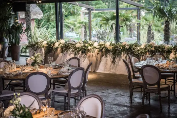 Elegant dining setup features round tables with white chairs, lush floral arrangements, and large glass windows showcasing greenery.