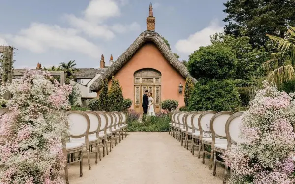 Charming outdoor ceremony space flanked by blooming flowers and elegant chairs, set against a quaint thatched cottage backdrop.