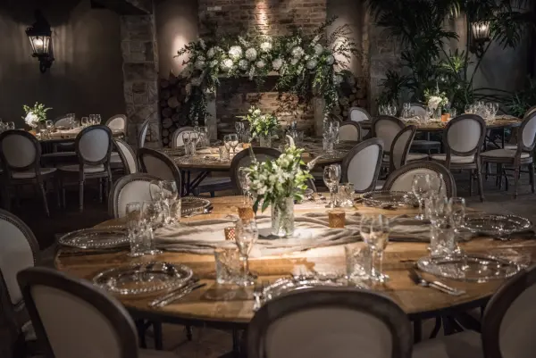 Intimate dining area featuring round wooden tables, elegant place settings, and lush floral arrangements against a rustic backdrop.