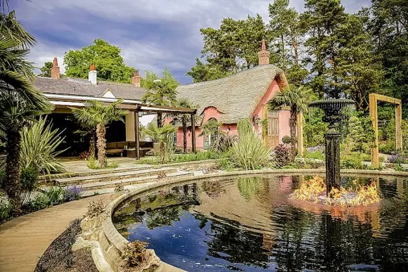 Charming wedding venue with a thatched roof, lush gardens, and a serene pond featuring a fountain and vibrant landscaping.