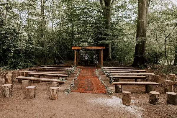 Charming outdoor ceremony space featuring rustic log seating, a vibrant aisle runner, and a natural wooden arch surrounded by lush greenery.