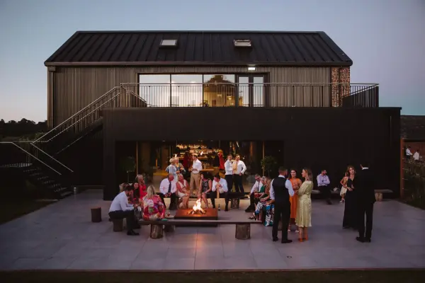 Guests gather around a cozy fire pit on a spacious patio, with a modern building and outdoor seating in the background, perfect for evening celebrations.