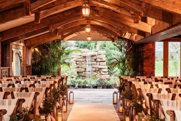 Charming indoor ceremony space with wooden beams, lush greenery, and a serene waterfall backdrop, framed by elegant seating.