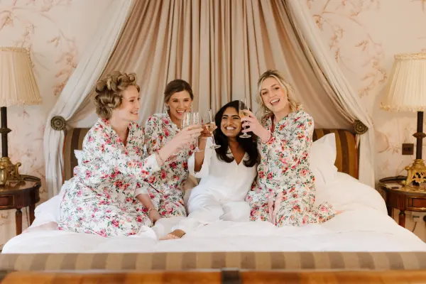 Bridesmaids in floral robes celebrate with champagne on a luxurious bed, featuring elegant decor and soft, neutral tones.